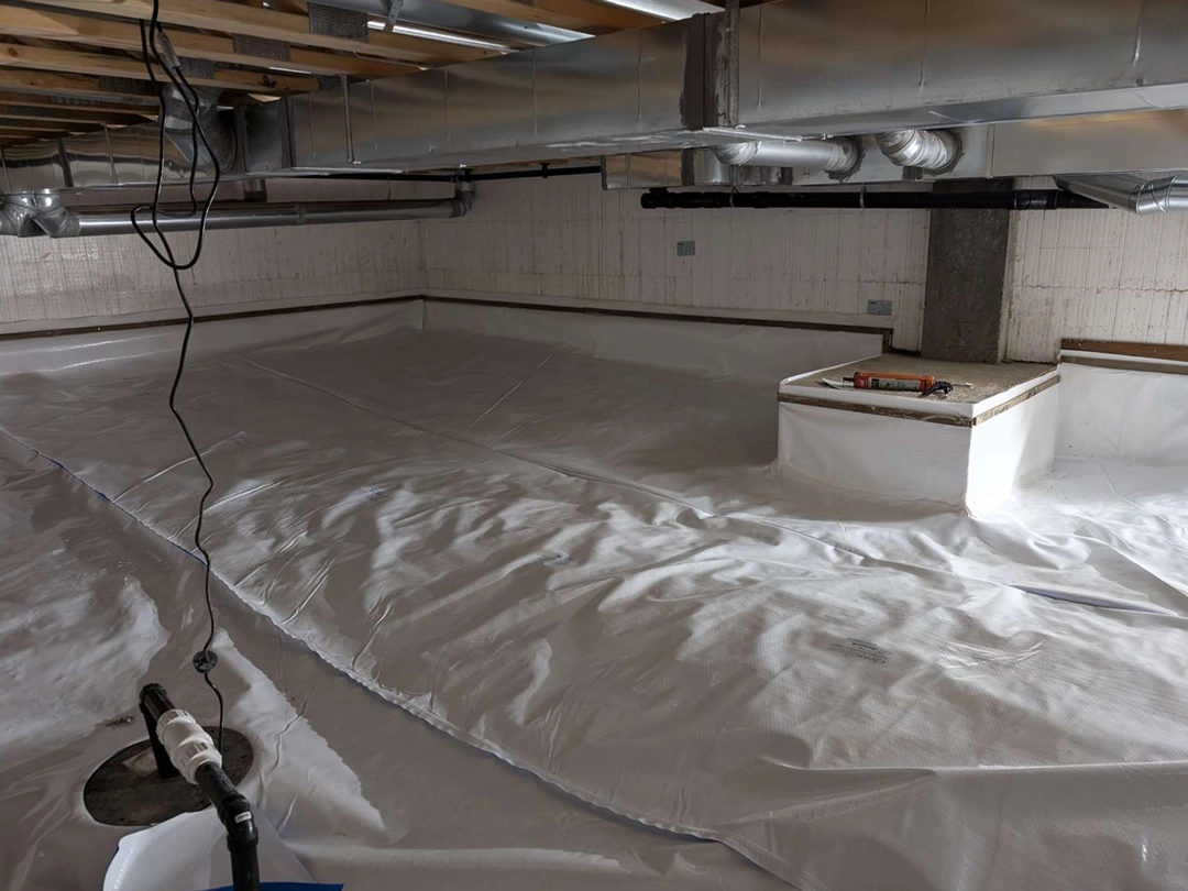 A basement with a white vapor barrier covering the floor, pipes overhead, and a small raised area with construction tools nearby