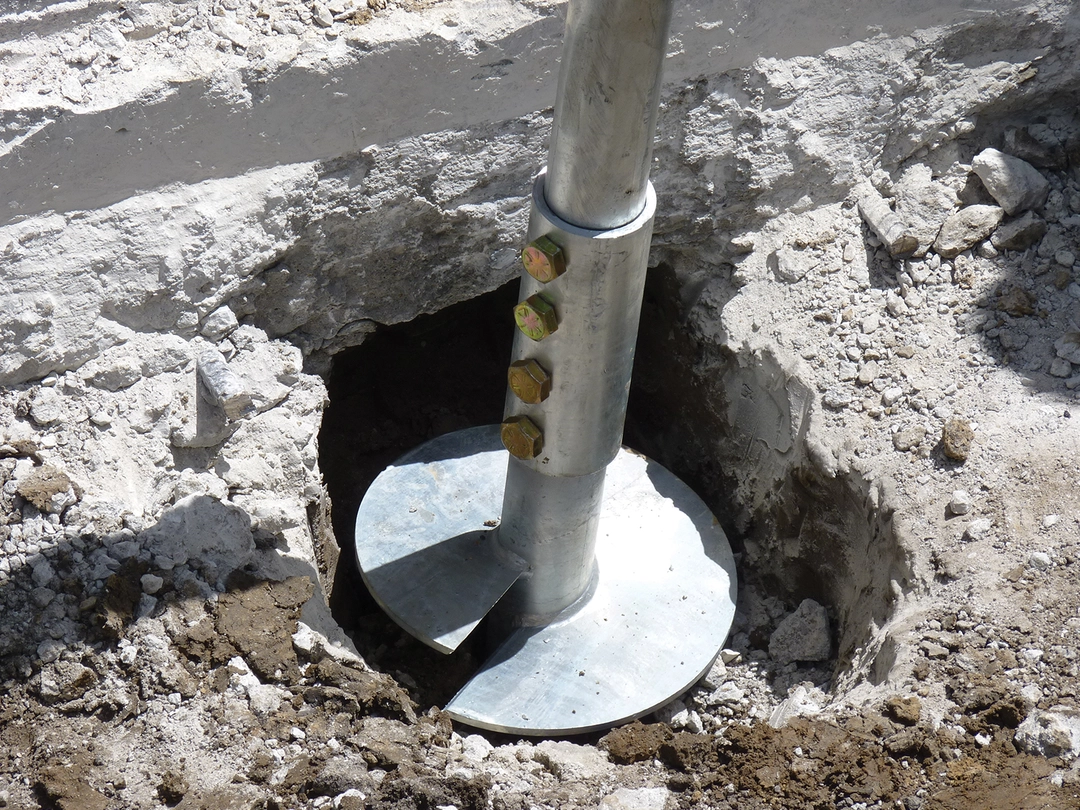 A large metal helical anchor installed in soil, with visible bolts and surrounding excavated earth