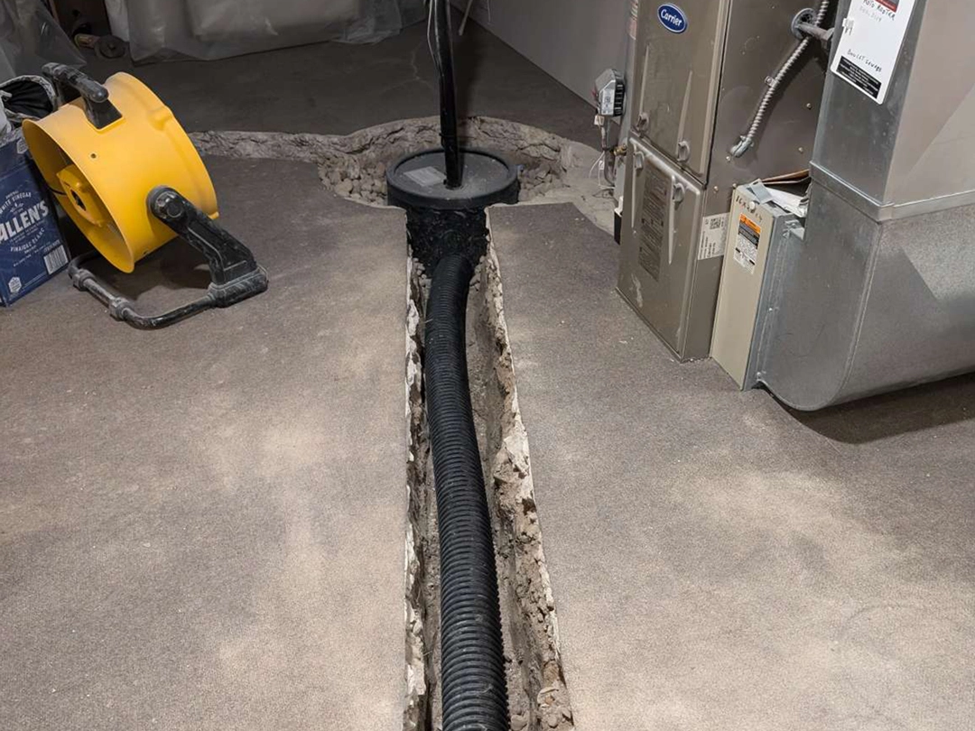 A basement with a cutout floor revealing a drainage trench, black piping, and a sump pump, alongside a yellow blower and HVAC unit
